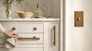 Person gently cleaning antique brass cabinet pulls and knobs with a soft cloth in a bright, modern kitchen.