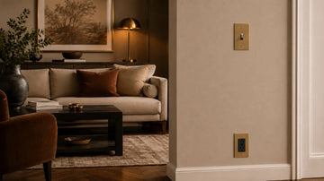 Brushed brass toggle light switch and matching black outlet installed at practical heights on a warm neutral living room wall with coordinated brass accents.