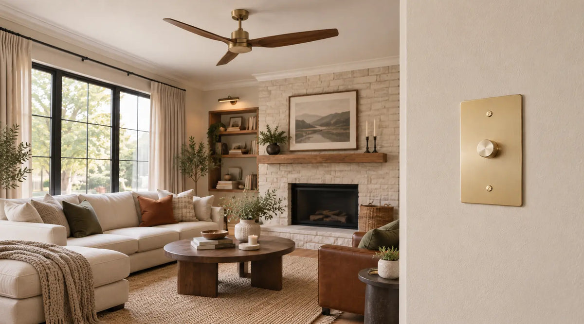 Brass fan control switch mounted on a living room wall with a ceiling fan visible in the background.