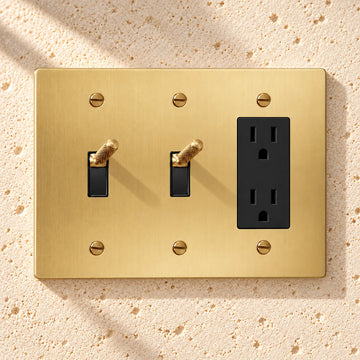 3-Gang Knurled Brass Toggle Light Switches & Duplex Outlet on sunlit pale limestone