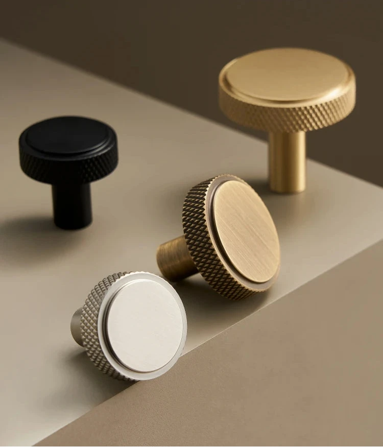 Four knurled brass knobs in black, gold, antique bronze and silver on a beige surface.