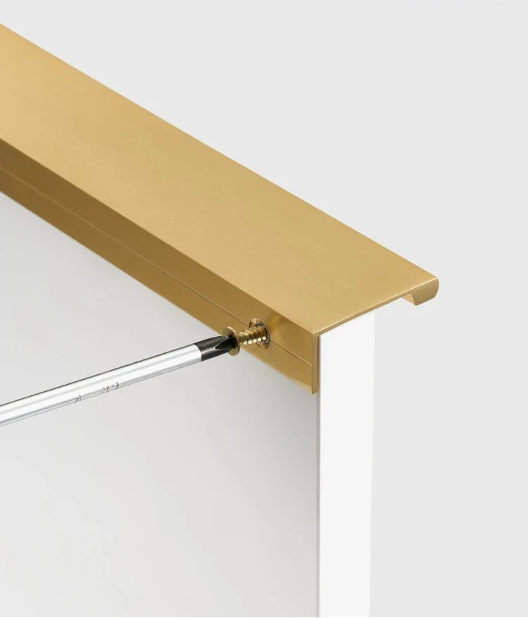 Gold brass edge pull being installed with a screwdriver on a white cupboard door.