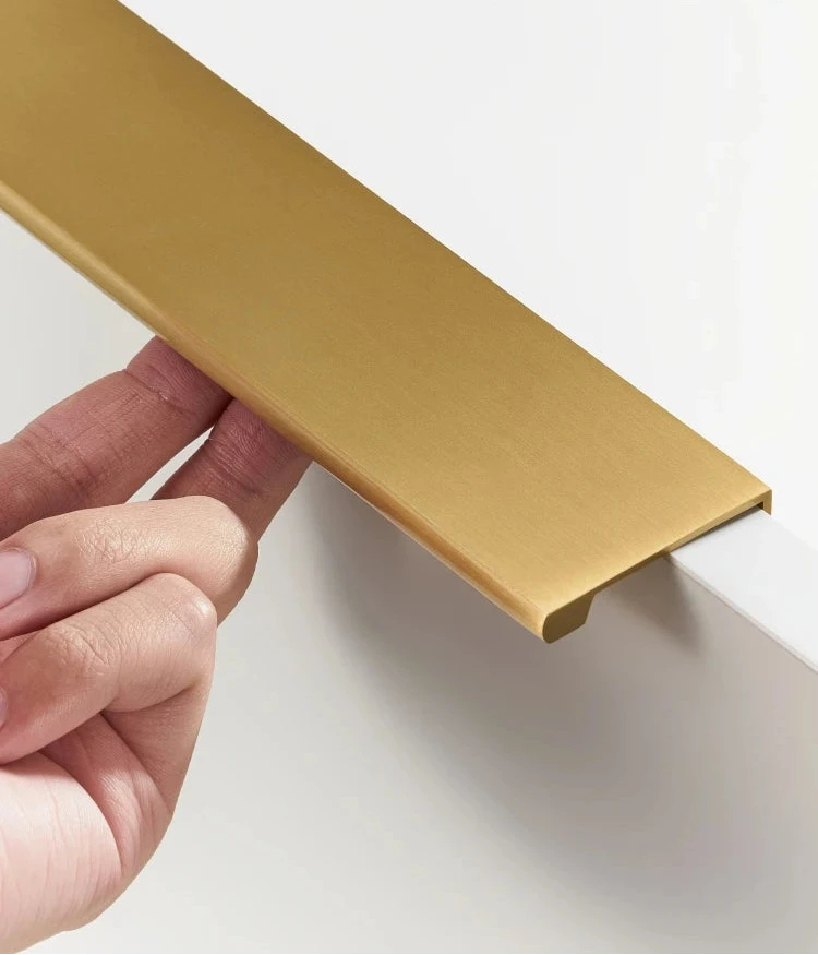 Hand pulling a strip of gold brass edge pull on a beige background.