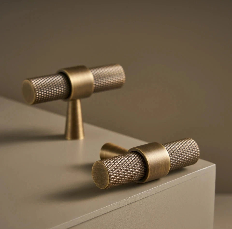 Two brass cabinet t-bars on a beige surface with a brown background.