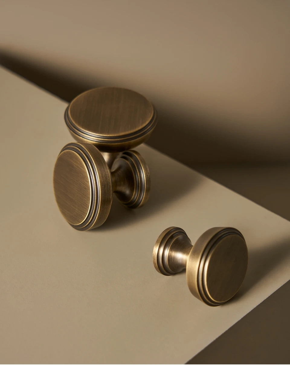 Two vintage brushed bronze cabinet knobs on a beige surface with a soft shadow. 