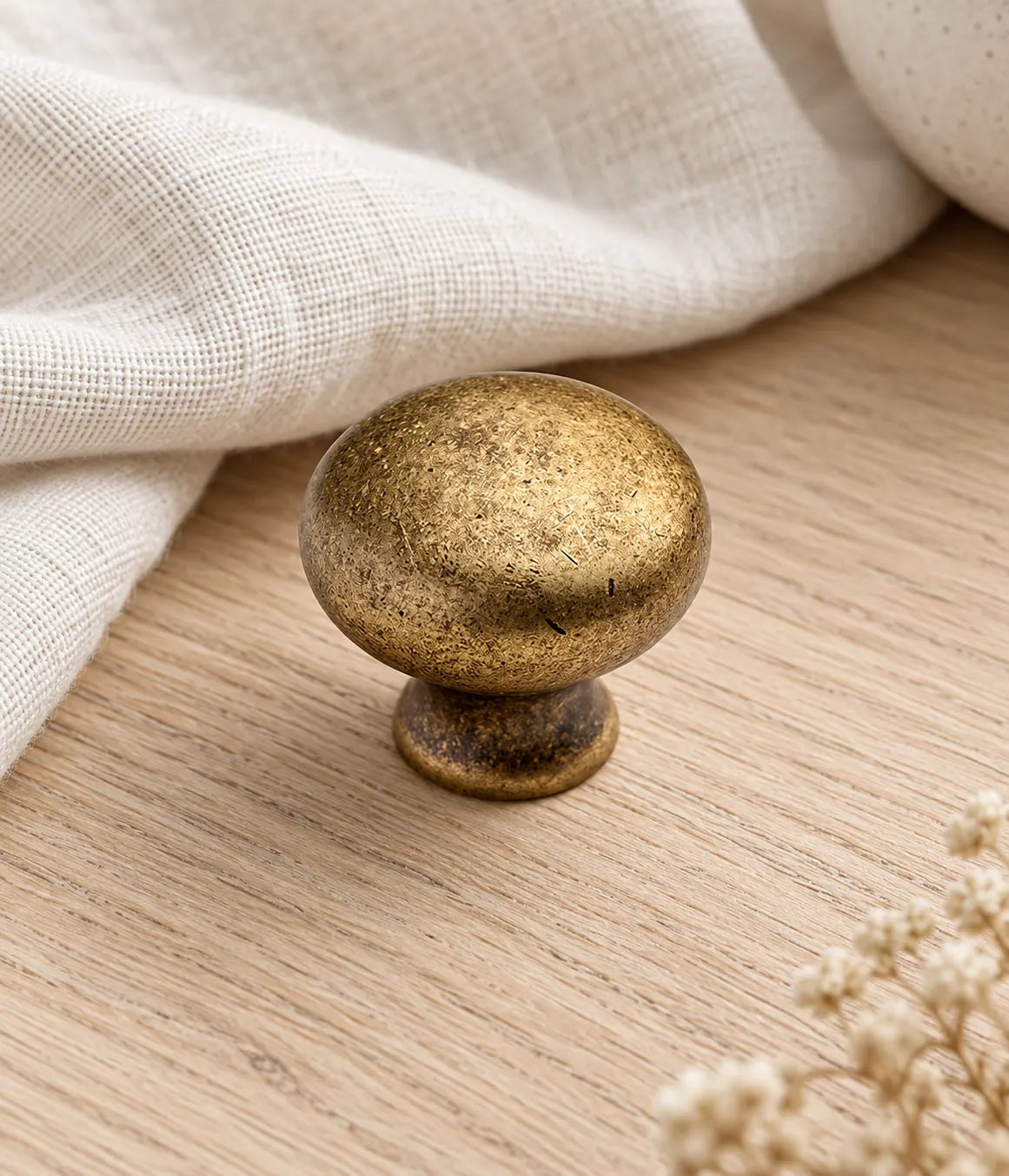 Distressed antique brass round cabinet knob on pale oak beside cream linen