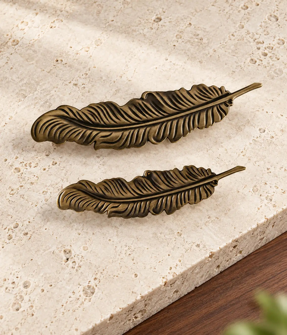 Two antique brass feather cabinet pulls arranged diagonally on warm limestone
