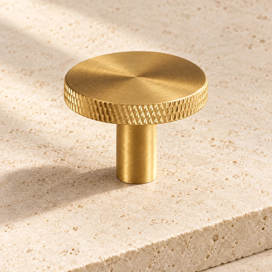 Brass cabinet knob with a flat circular face and diamond-knurled edge on pale limestone.