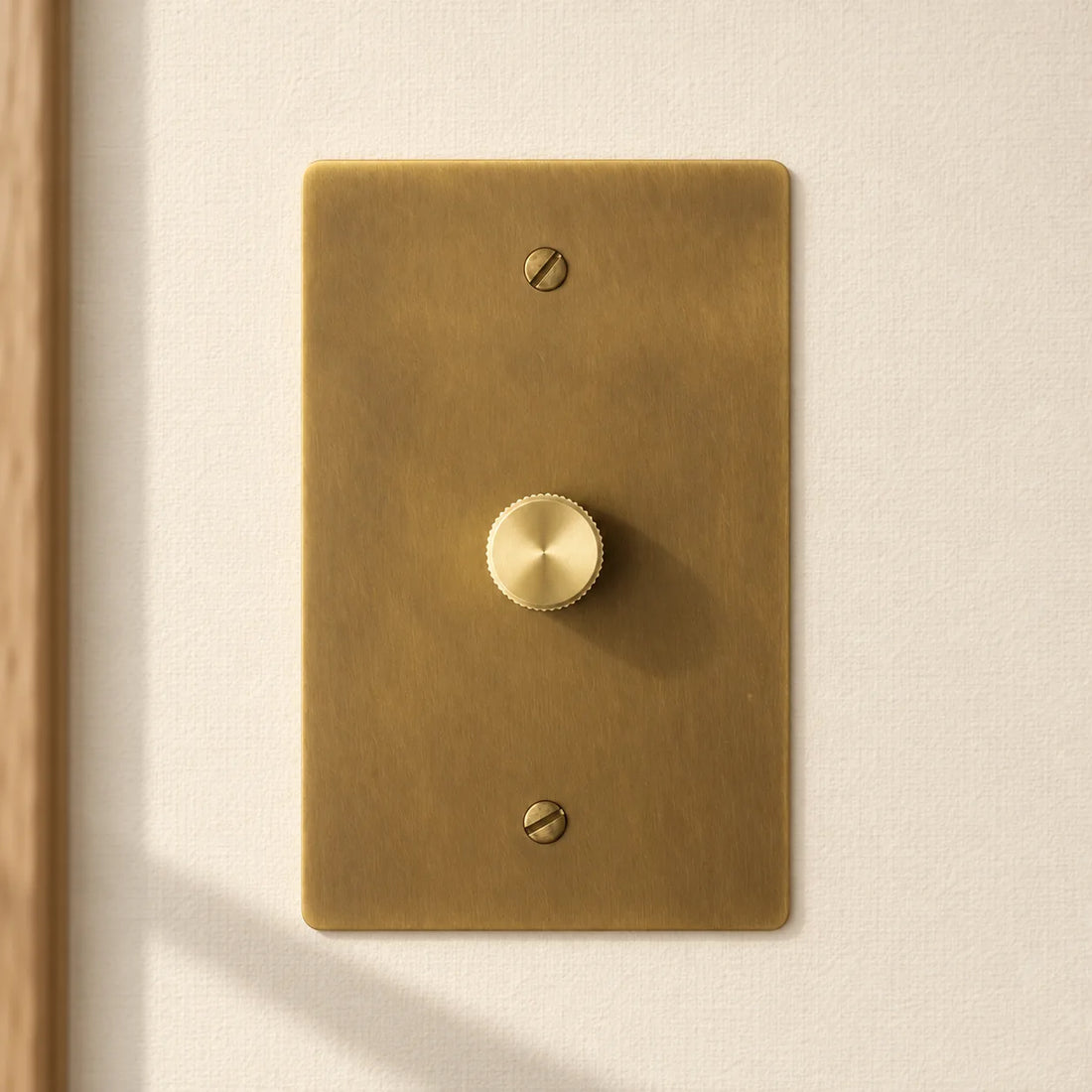 1-gang antique brass rotary dimmer switch with one matching knurled knob and slotted screws, installed on a warm cream textured wall in natural sunlight.