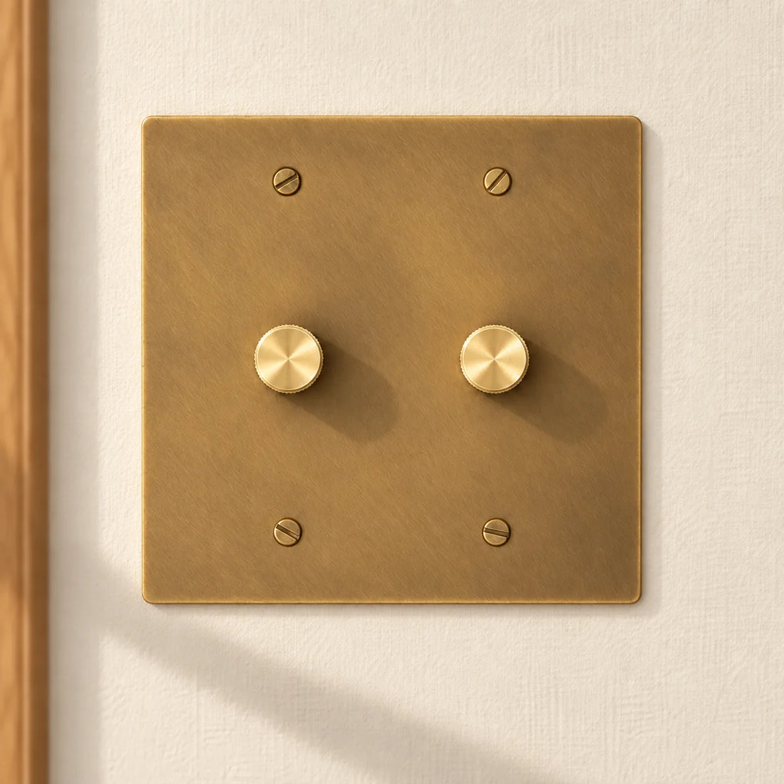 2-gang antique brass rotary dimmer switch with 2 matching knurled knobs and slotted screws, installed on a warm cream textured wall in natural sunlight.