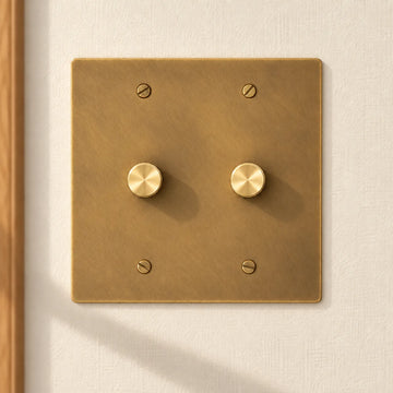 2-gang antique brass rotary dimmer switch with 2 matching knurled knobs and slotted screws, installed on a warm cream textured wall in natural sunlight.