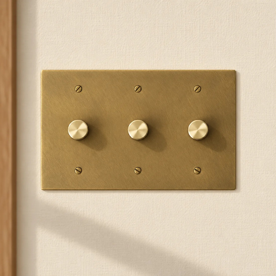 3-gang antique brass rotary dimmer switch with 3 matching knurled knobs and slotted screws, installed on a warm cream textured wall in natural sunlight.