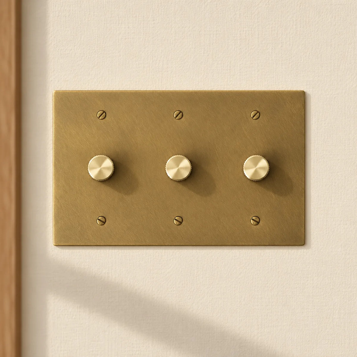 3-gang antique brass rotary dimmer switch with 3 matching knurled knobs and slotted screws, installed on a warm cream textured wall in natural sunlight.