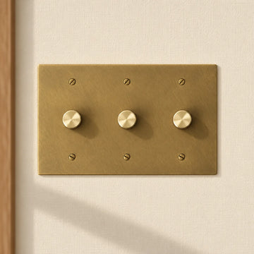 3-gang antique brass rotary dimmer switch with 3 matching knurled knobs and slotted screws, installed on a warm cream textured wall in natural sunlight.