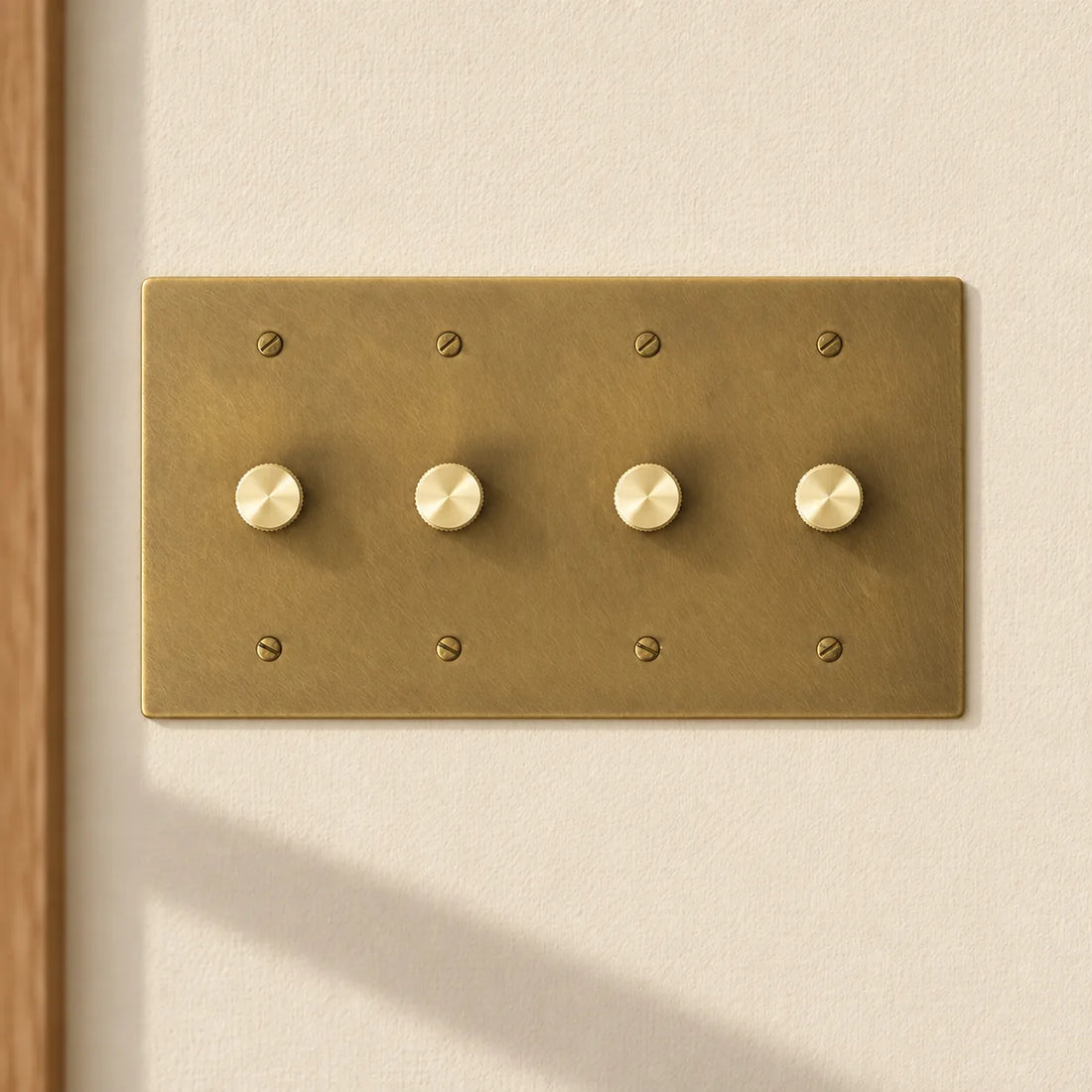 4-gang antique brass rotary dimmer switch with 4 matching knurled knobs and slotted screws, installed on a warm cream textured wall in natural sunlight.