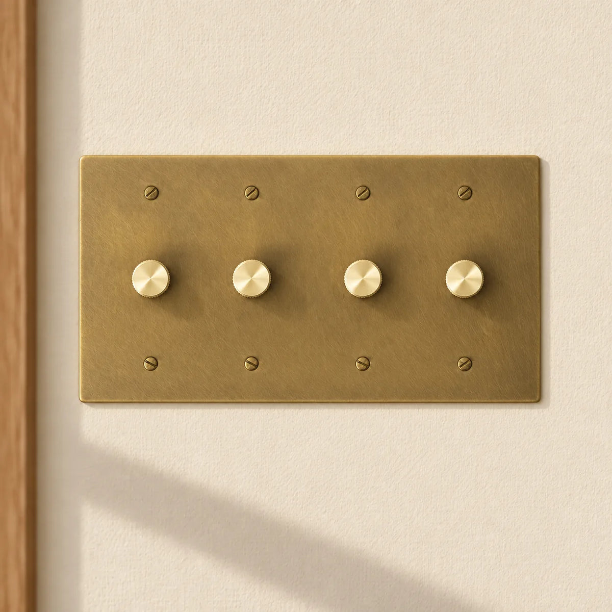 4-gang antique brass rotary dimmer switch with 4 matching knurled knobs and slotted screws, installed on a warm cream textured wall in natural sunlight.