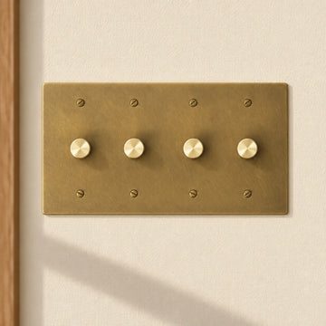 4-gang antique brass rotary dimmer switch with 4 matching knurled knobs and slotted screws, installed on a warm cream textured wall in natural sunlight.