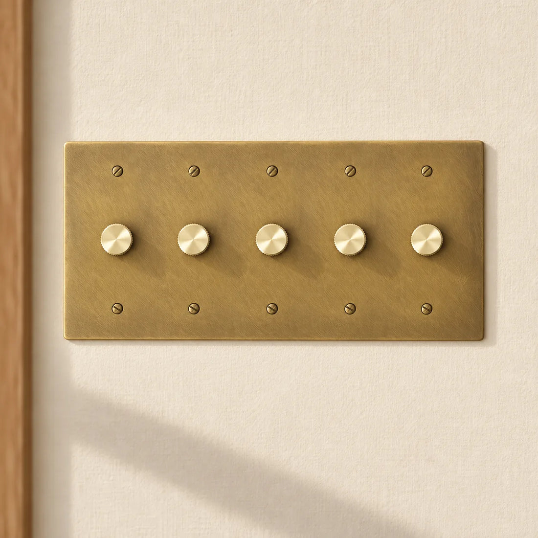5-gang antique brass rotary dimmer switch with 5 matching knurled knobs and slotted screws, installed on a warm cream textured wall in natural sunlight.