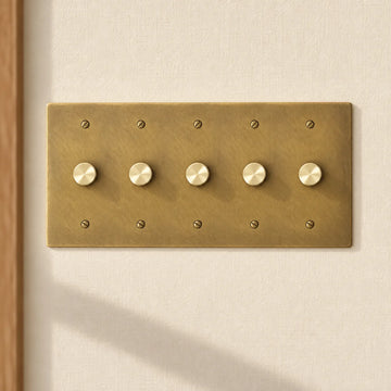 5-gang antique brass rotary dimmer switch with 5 matching knurled knobs and slotted screws, installed on a warm cream textured wall in natural sunlight.