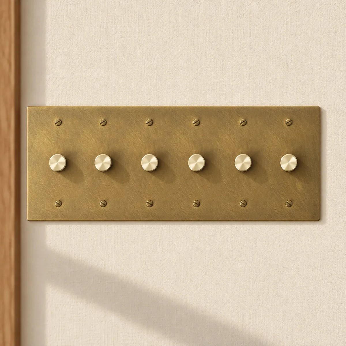 6-gang antique brass rotary dimmer switch with 6 matching knurled knobs and slotted screws, installed on a warm cream textured wall in natural sunlight.