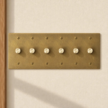 6-gang antique brass rotary dimmer switch with 6 matching knurled knobs and slotted screws, installed on a warm cream textured wall in natural sunlight.