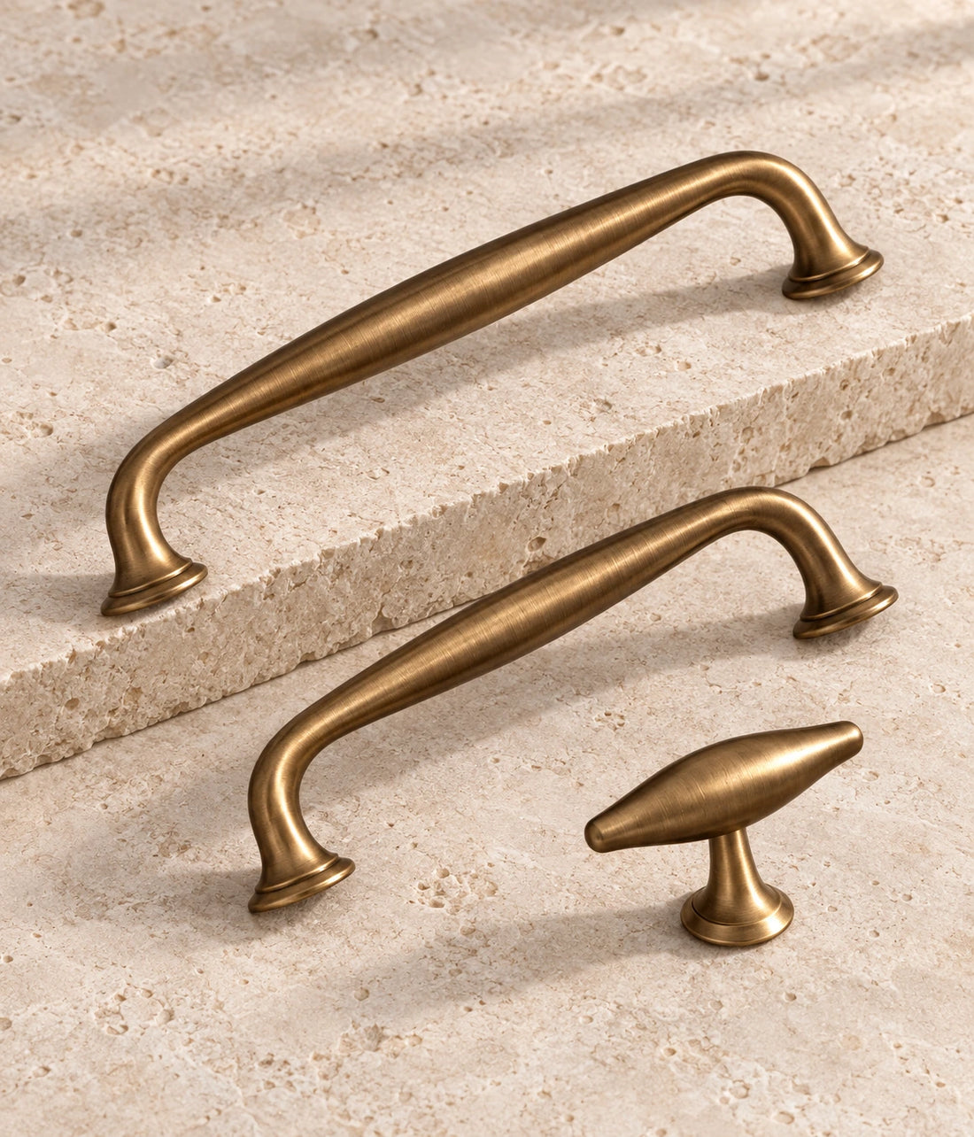Antique Brass Arched Cabinet Pulls & Knobs arranged on warm limestone, showing the antique brushed brass finish