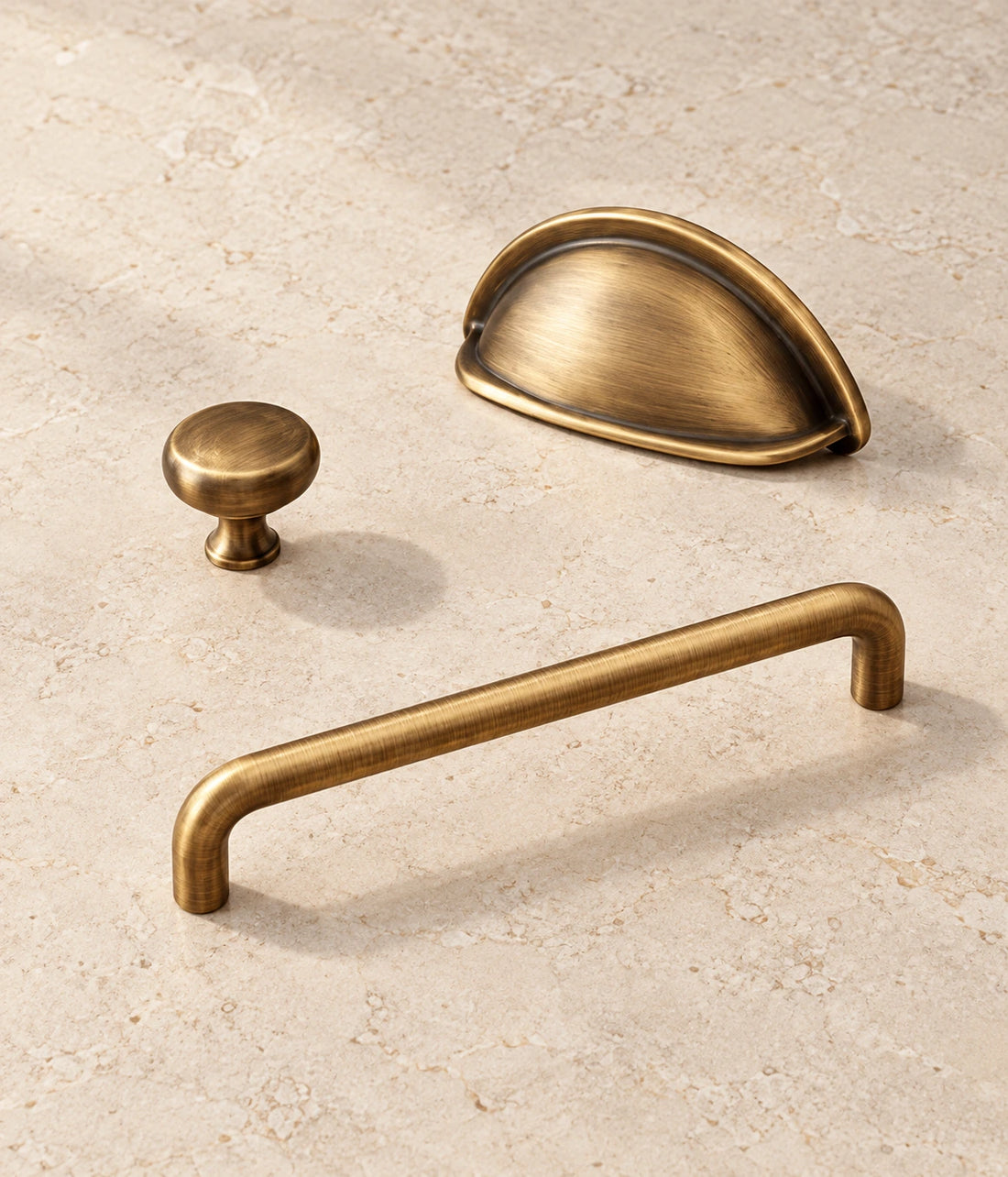Antique Brass Rounded Cabinet Pulls, Cup Pull & Knob arranged on pale limestone, showing the brushed brass finish and hardware details